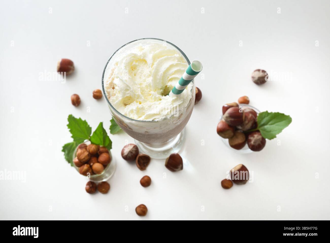 Glas mit Haselnuss-Smoothie mit Creme und grünem Strohhalm auf weißem Tisch mit Schüsseln voller Haselnüsse und Blättern auf weißem Tisch. Oben Stockfoto