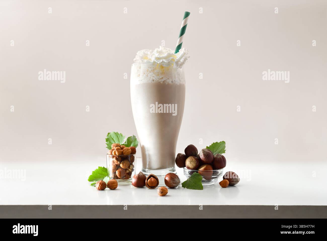 Glas mit Haselnuss-Smoothie mit Creme und grünem Stroh auf weißem Tisch mit Schüsseln voller Haselnüsse und Blätter auf isoliertem hellen Hintergrund. Frontscheibe Stockfoto