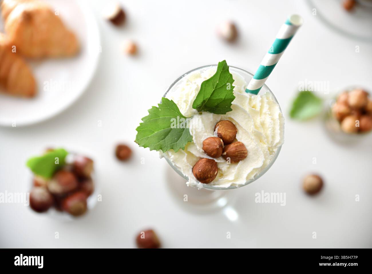 Detail aus Glas mit Haselnussmilkshake mit Creme und grünem Stroh, dekoriert mit Haselnüssen und Blättern auf einem weißen Tisch mit Schüsseln voller Früchte und Stockfoto