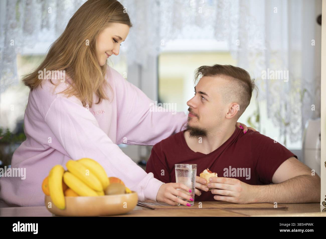 Eine lächelnde blonde Frau mit einem rosa Sweatshirt steht in der Küche. Ein Mann mit Bart sitzt am Tisch und isst ein Sandwich Stockfoto