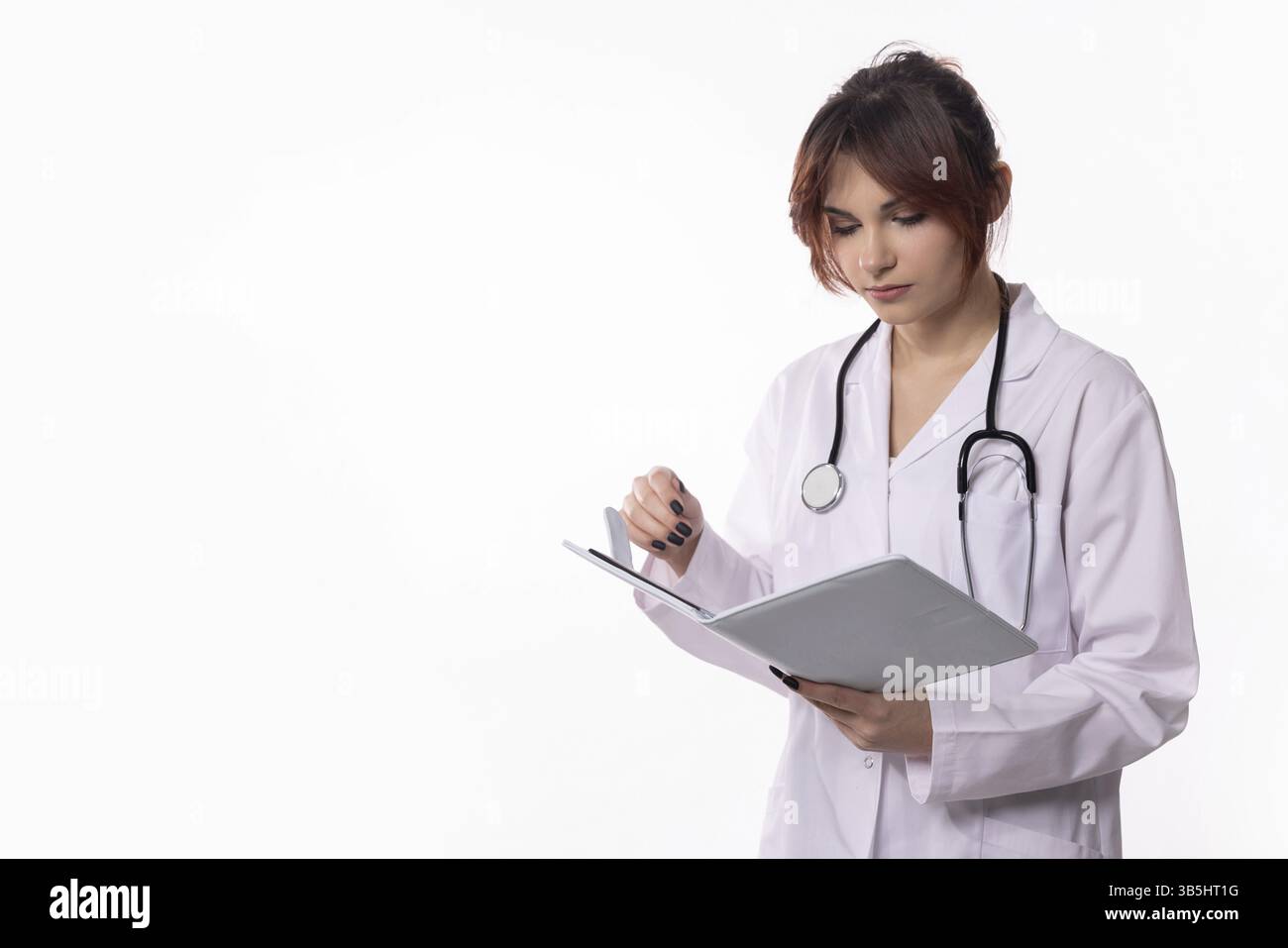 Eine hingebungsvolle Ärztin untersucht Patientennotizen in einem knusprigen weißen Mantel, mit einem Stethoskop um den Hals Stockfoto