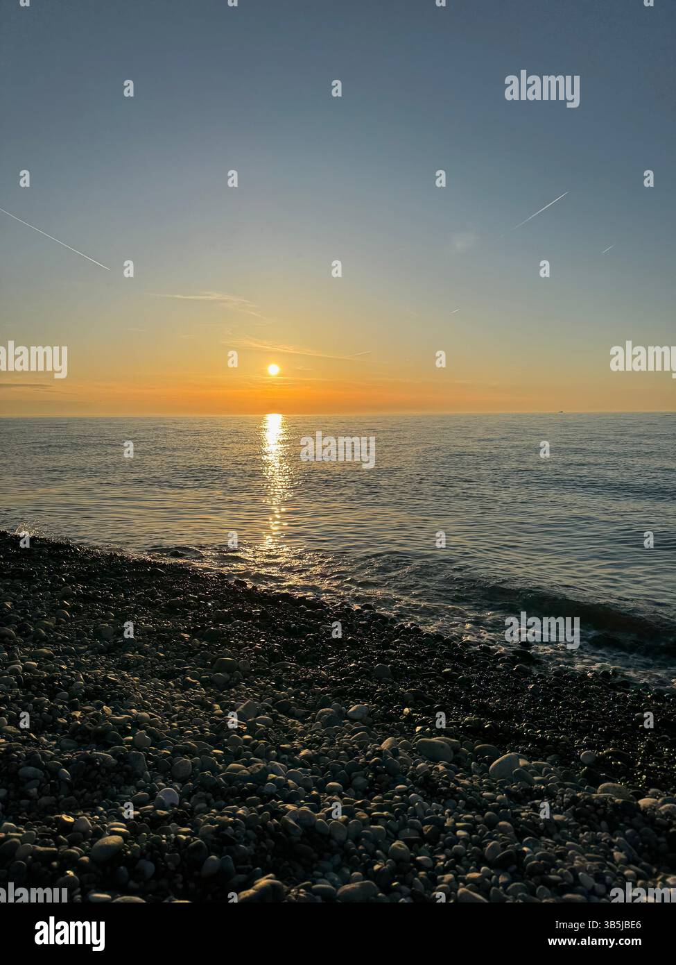 Kieselstrand bei Sonnenuntergang mit ruhigem Meer und Sonnenspiegelung auf dem Wasser. Sanfte Farben, natürliches Licht, friedliche Küstenlandschaft, keine Leute, Abendstimmung. - Smartphone-aufgenommenes Stockfoto