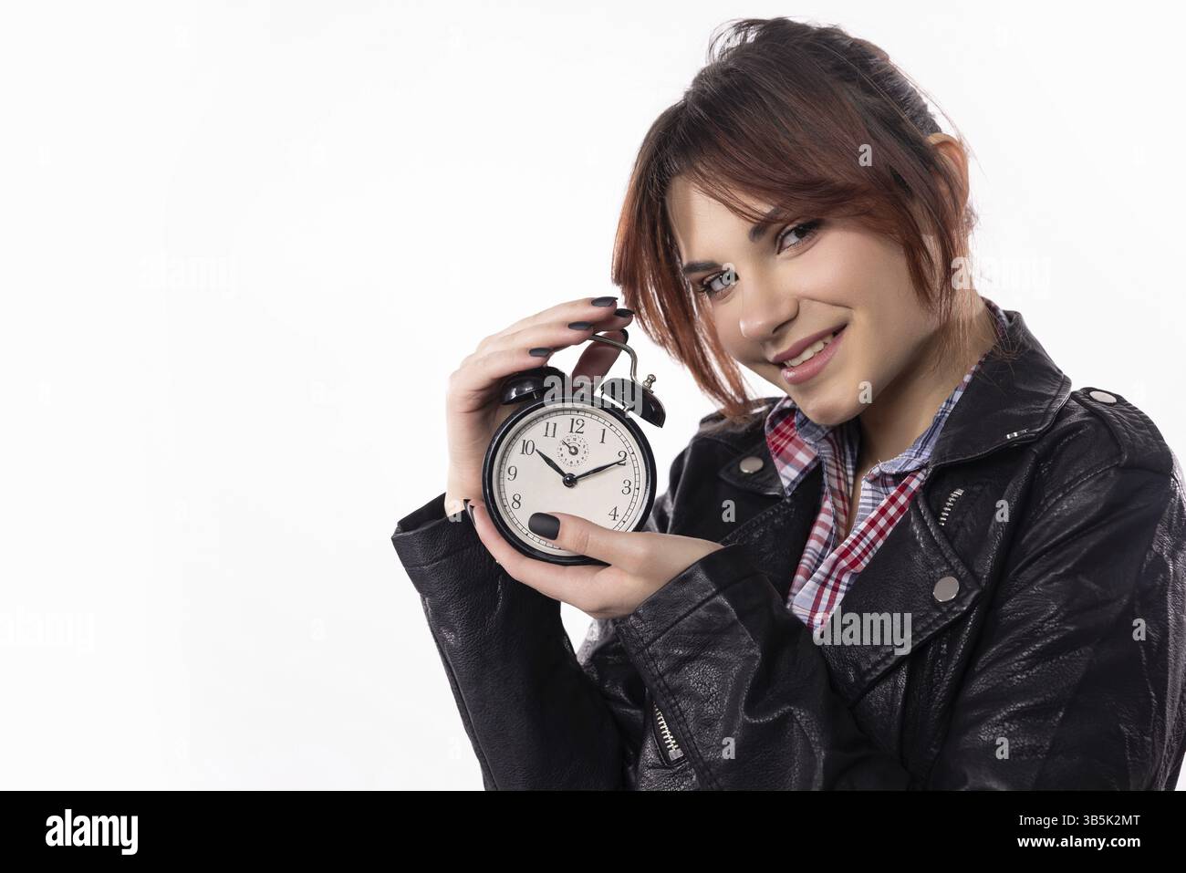 Eine stilvolle junge Frau lächelt breit, während sie selbstbewusst einen Wecker hält und ein freudiges Gefühl der Dringlichkeit zeigt Stockfoto