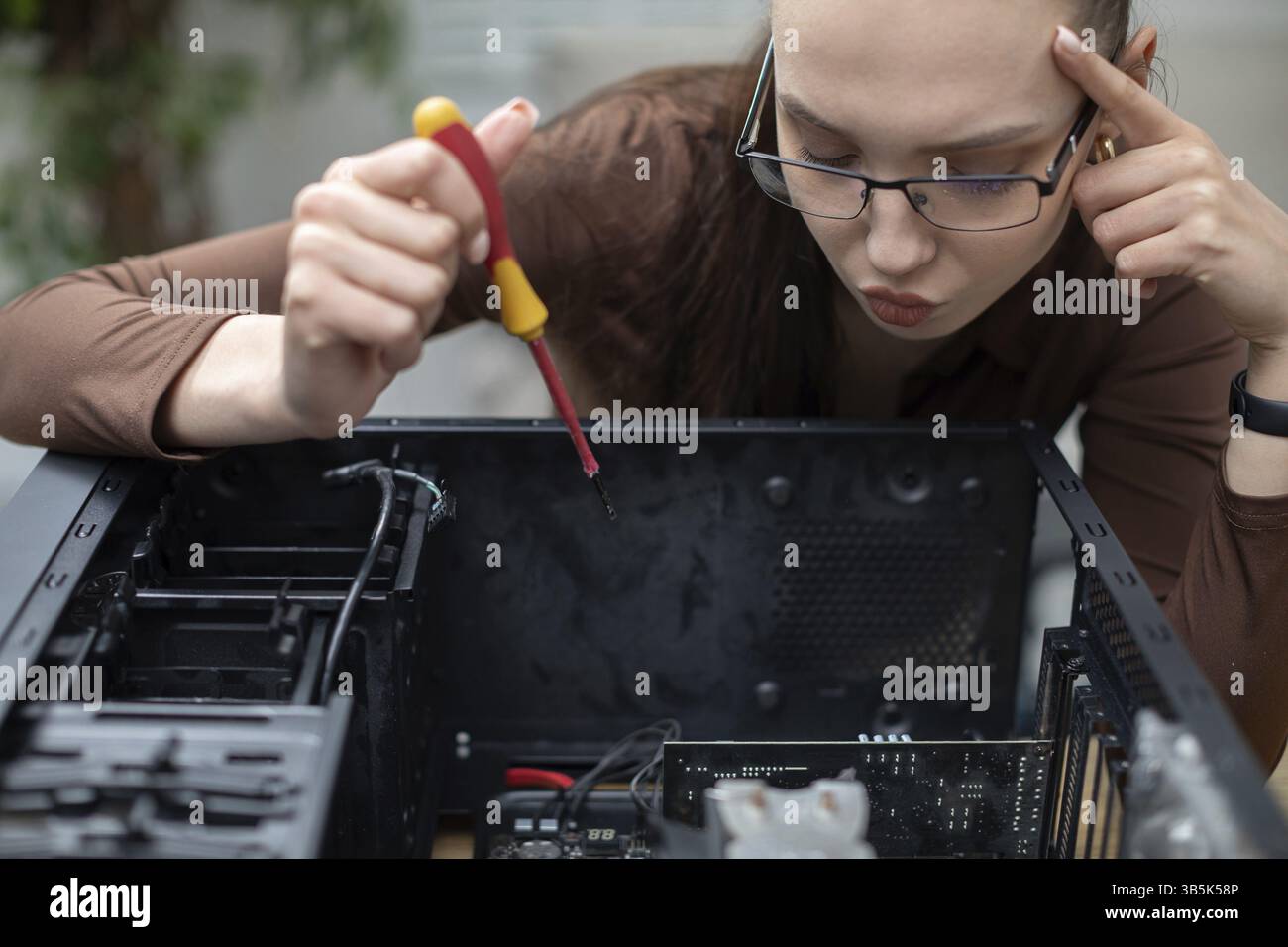 Eine Frau lehnt sich über einen Computer. Die Frau hat einen nachdenklichen Ausdruck im Gesicht. Der Computer wird auseinandergenommen. Die Frau hält einen Schraubenzieher in ihrem Ha Stockfoto