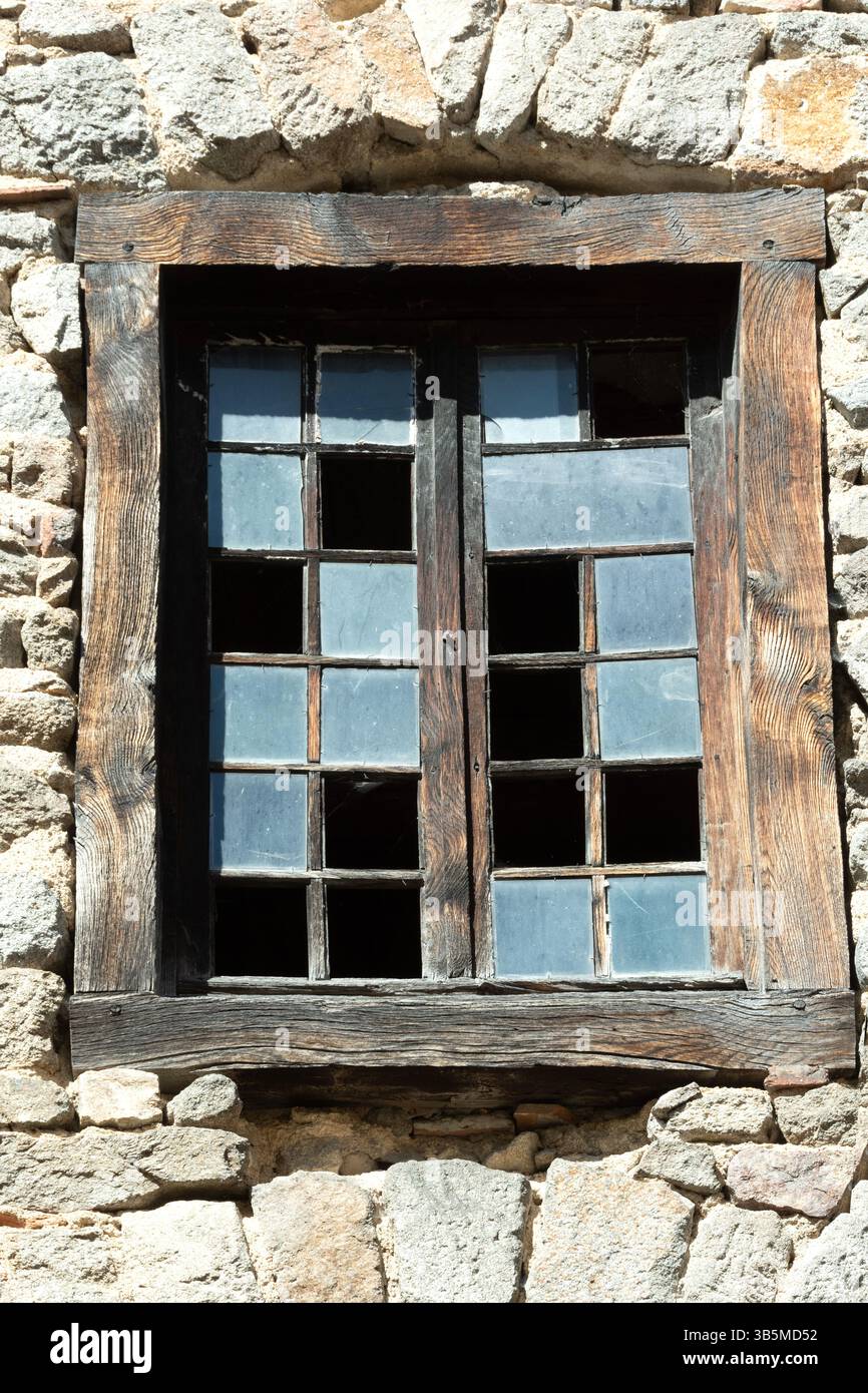 Ein verwittertes Holzfenster mit mehreren gebrochenen Glasscheiben ist in eine rustikale Steinmauer eingefasst. Das Sonnenlicht wirft Schatten und hebt die Texturen des hervor Stockfoto