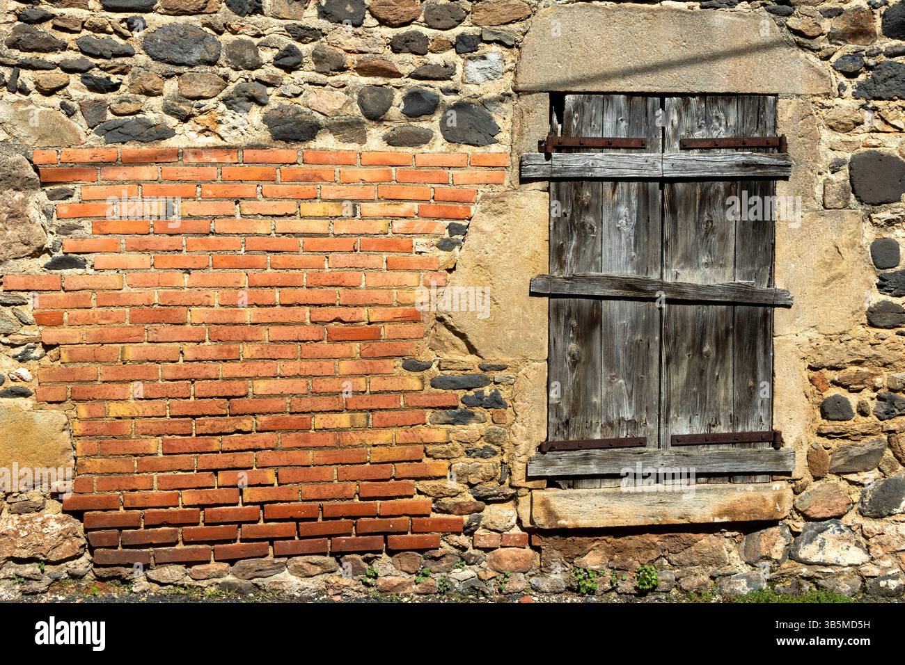 Eine rustikale Wand kombiniert strukturierte rote Ziegelsteine und alte Steine, ergänzt durch Holzläden. Sonnenlicht wirft Schatten. Frankreich Stockfoto