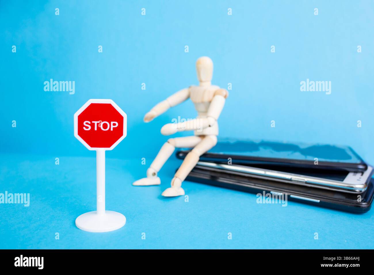 Hölzerne Schaufensterpuppe, die auf einem Smartphone-Stapel sitzt, mit einem Stoppschild auf blauem Hintergrund Stockfoto
