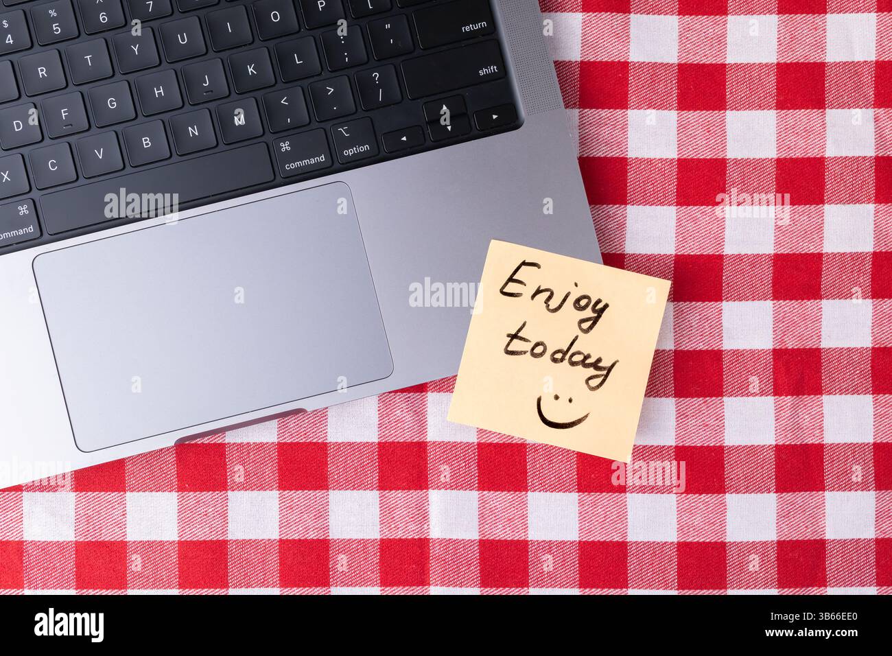 Haftnotiz mit „heute genießen“-Text und Smiley-Gesicht neben einem Laptop auf einer rot-weiß karierten Oberfläche, die eine positive Erinnerung an darstellt Stockfoto