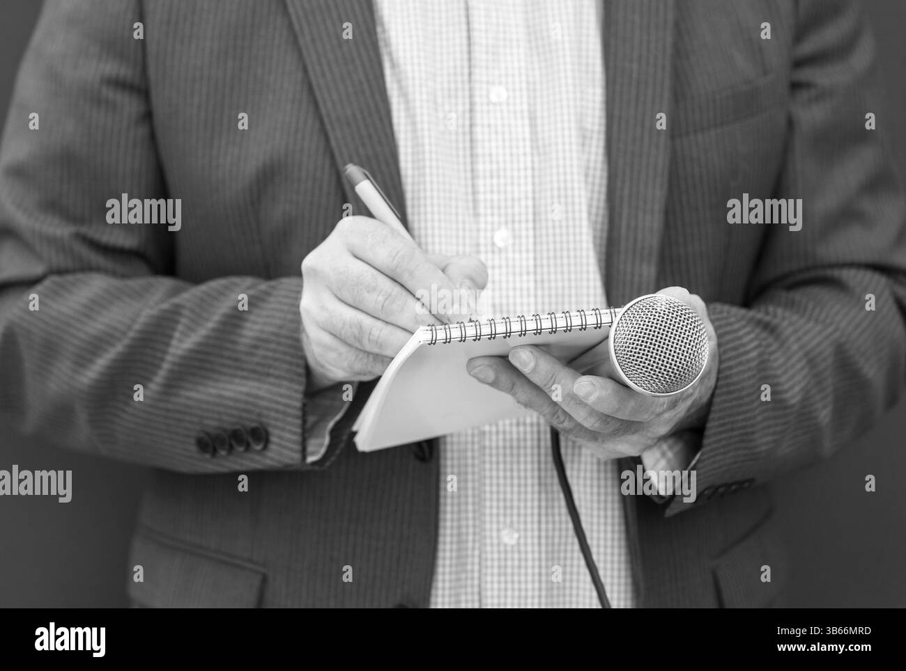 Ein professioneller Journalist, der einen Anzug trägt, nimmt während eines wichtigen Medieninterviews sorgfältig handschriftliche Notizen in einen Notizblock, während er ein Mikrofon hält Stockfoto