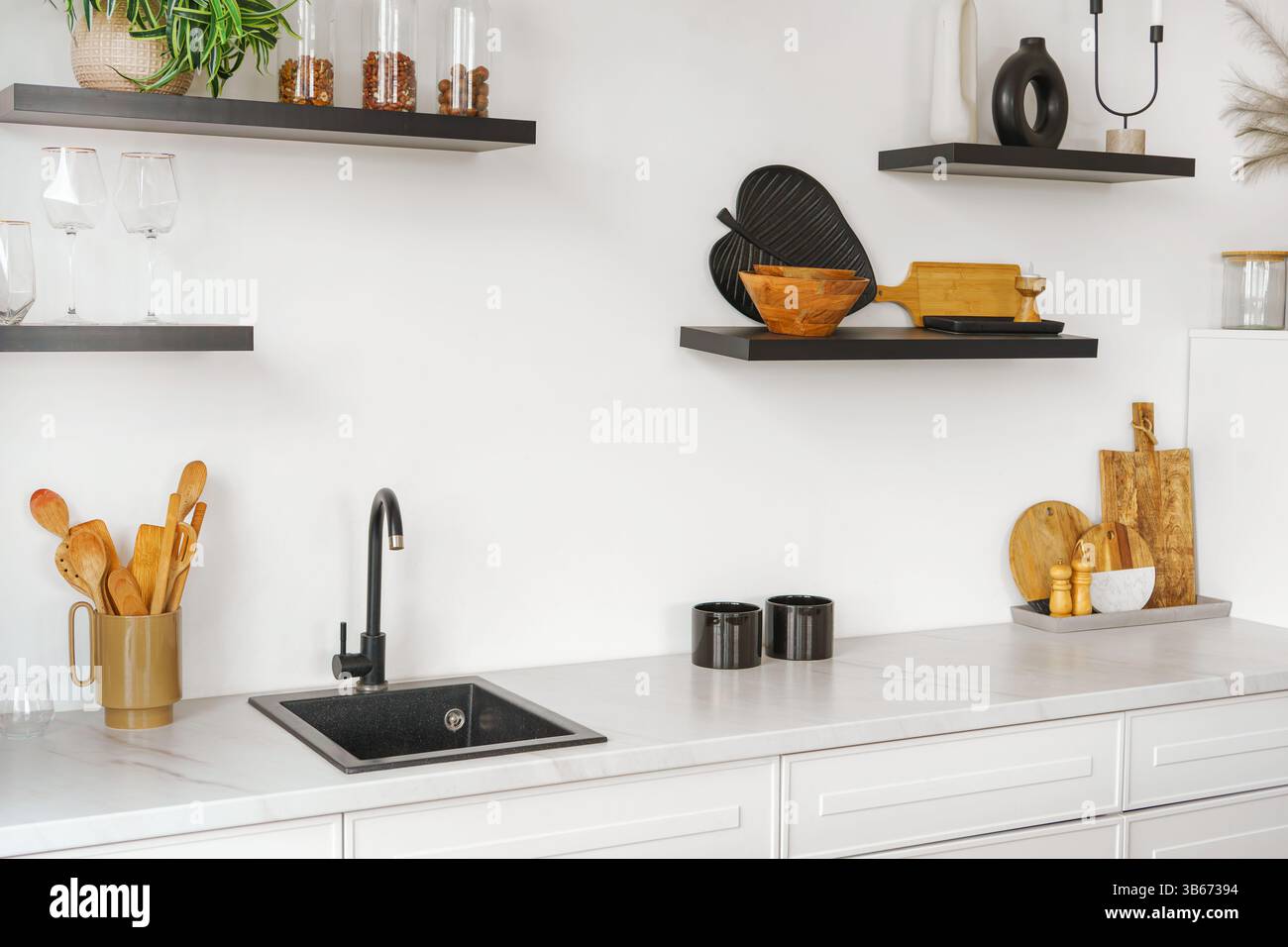 Weiße Küche mit Arbeitsplatte und schwarzem Wasserhahn auf weißem Wandhintergrund mit Holzregalen. Regale mit Utensilien, Küchenschrank und Einrichtung. Design in Stockfoto