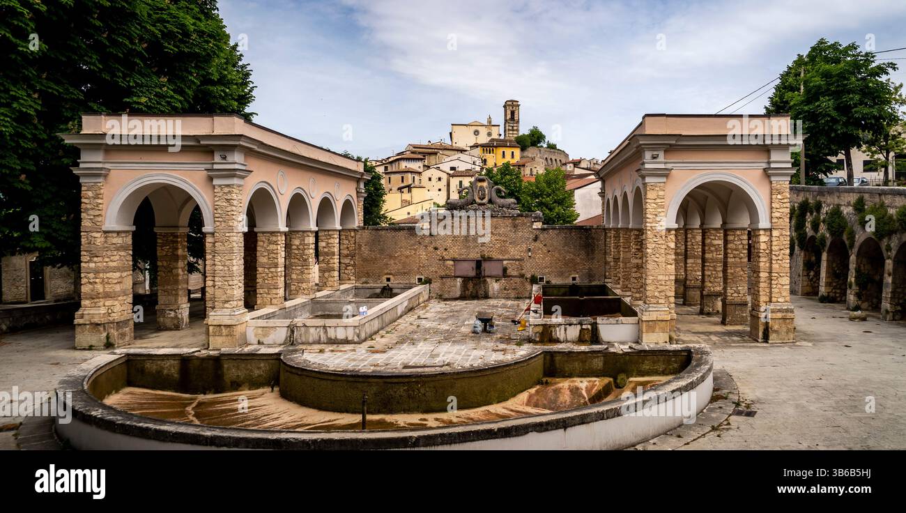 Ein historischer Brunnen, umgeben von Steinbögen und Grün, mit einem Dorf im Hintergrund. Die Szene fängt eine Mischung aus Architektur und Natur ein, Stockfoto