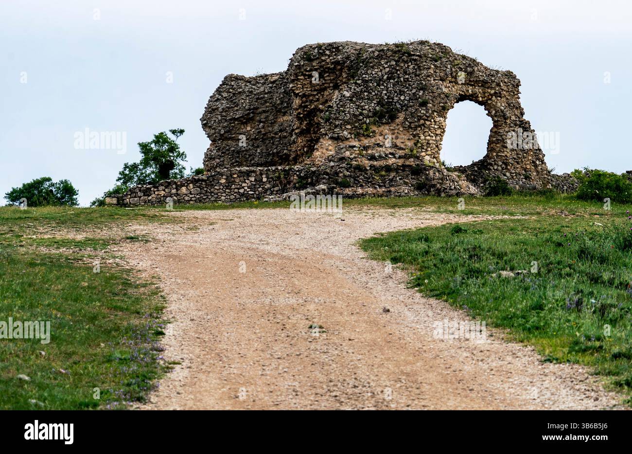 Eine verwitterte Steinruine mit einem Bogengang, vor einer grasbewachsenen Landschaft. Ein Schotterweg führt zum Bauwerk, umgeben von Grün und Bäumen unter Stockfoto