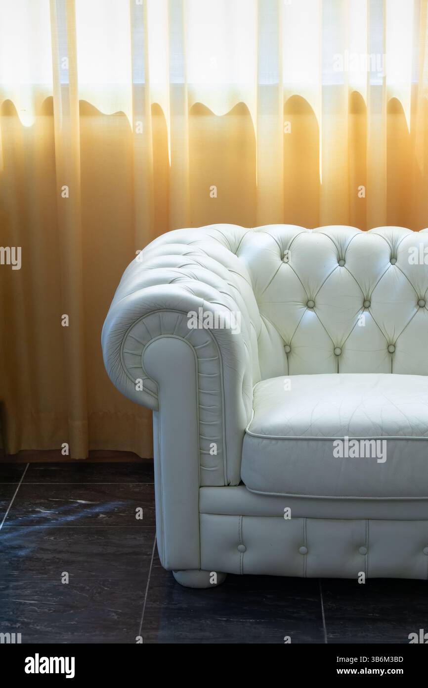 Weißes chesterfield-Ledersofa in einem Luxushotel in der Nähe eines Fensters mit eleganten gelben Vorhängen Stockfoto