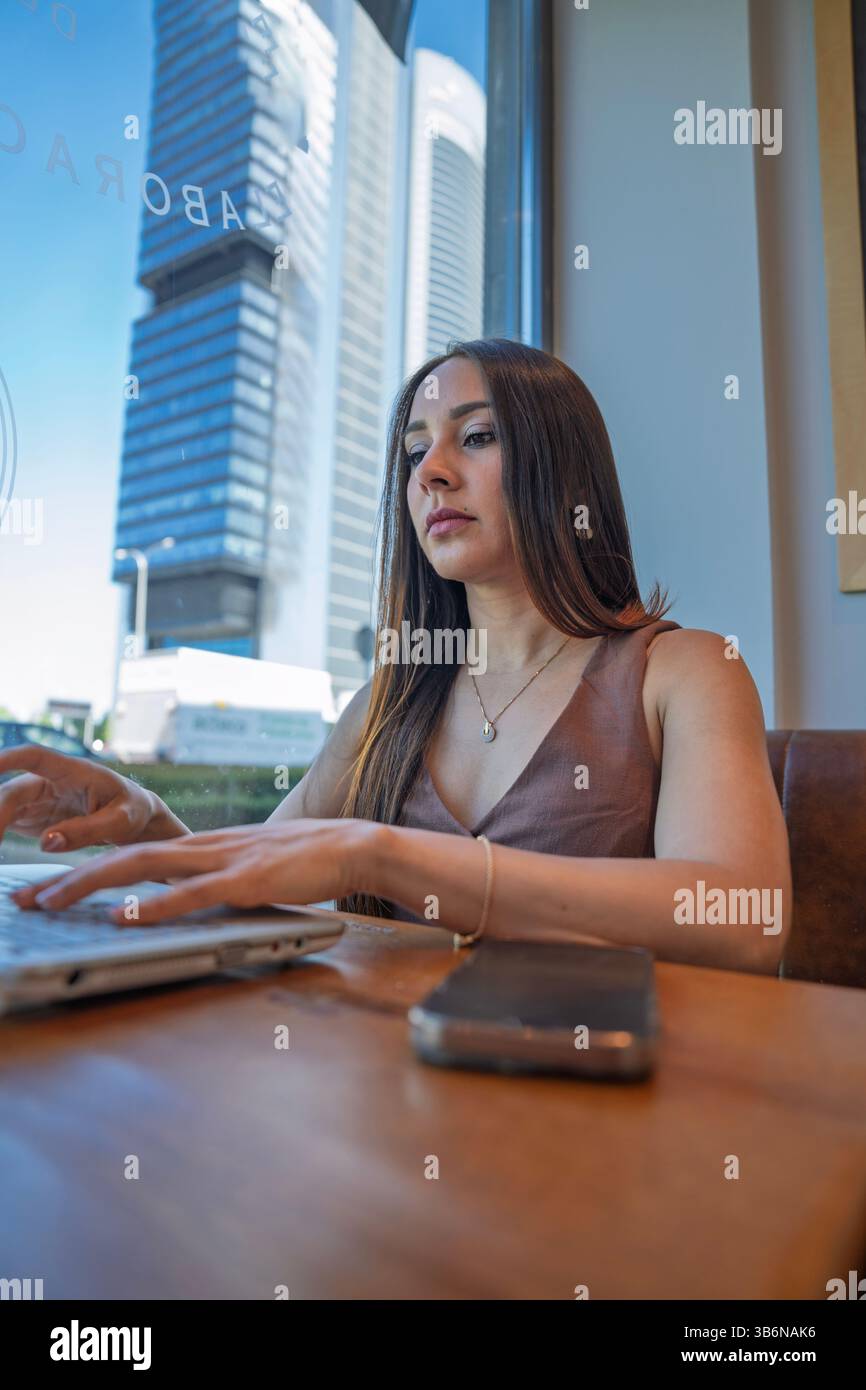 Junger Unternehmer mit Laptop in einem Café, der von natürlichem Licht und Stadtlandschaft profitiert Stockfoto