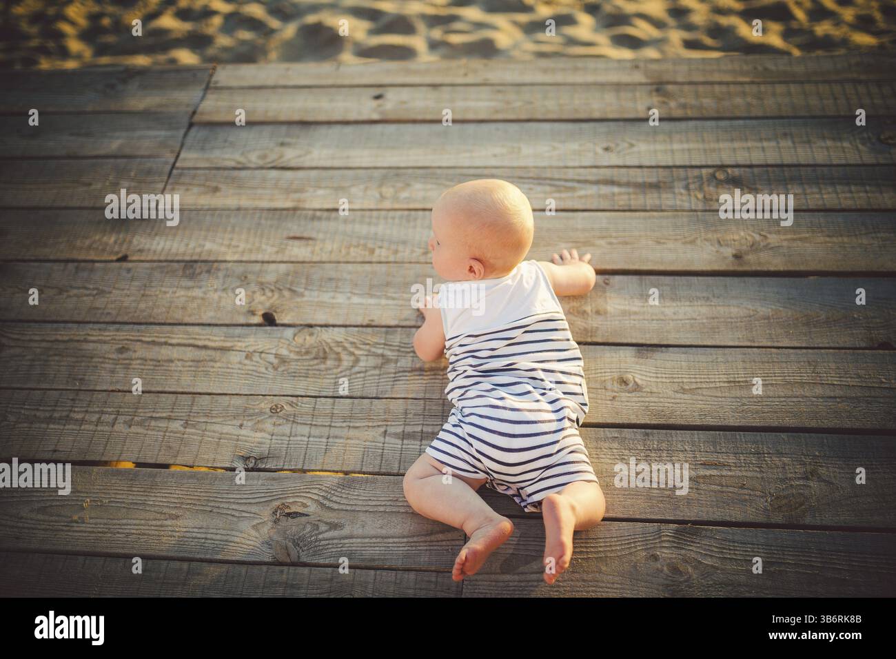 Neugeborene Kind ein Junge, ein Jahr alt, liegt auf seinem Bauch auf ein hölzernes Dock, einem Pier in gestreifter Kleidung, eine Sicht von oben im Sommer eine komplet bei Sonnenuntergang Stockfoto