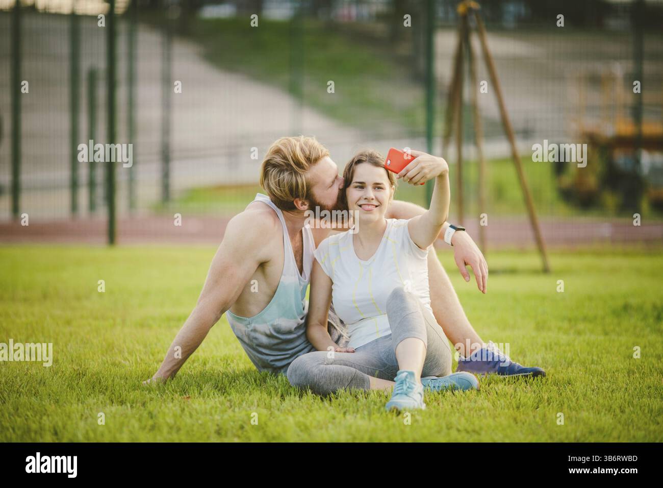 Sport und Technik. Junge in der Liebe heterosexuellen Kaukasischen paar Ausruhen nach Training draußen im Park auf Rasen, grünes Gras sitzen in Umarmung und m Stockfoto