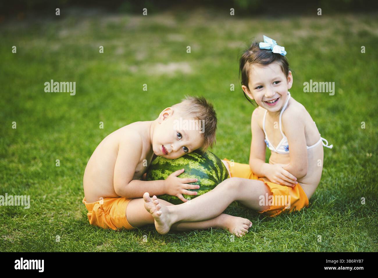 Zwei Kinder, kaukasische Bruder und Schwester, auf grünem Gras im Hinterhof des Hauses sitzen und umarmen Big leckere Süße Wassermelonen Berry, grün gestreiften Co Stockfoto