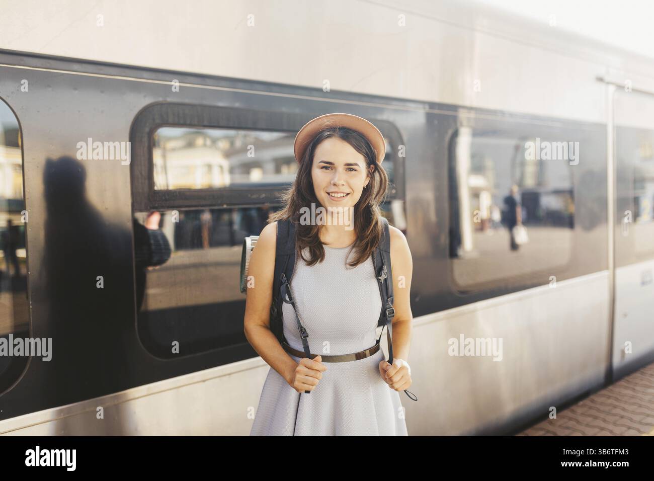 Thema Eisenbahn und Reisen. Portrait der jungen kaukasischen Frau mit Zahnlächeln, die am Bahnhofshintergrund mit Rucksack und Ausrüstung steht Stockfoto