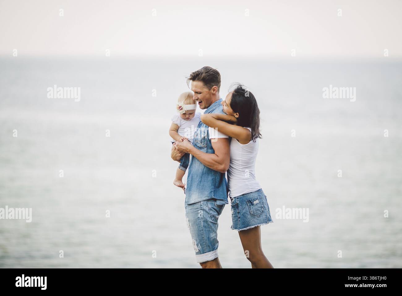 Familienurlaub in der Natur. Drei, Mutter, Vater, Tochter ein Jahr ständigen mit Rücken auf einer Klippe mit Blick auf das Meer. Mann hält Hild in Arme, Frau umarmen Hus Stockfoto