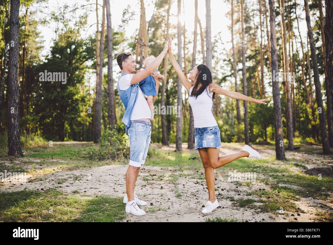 Familie Mama, Papa und Tochter sitzt bei Papa auf den Schultern, und Eltern Kuss auf die Natur im Wald im Sommer bei Sonnenuntergang Stockfoto