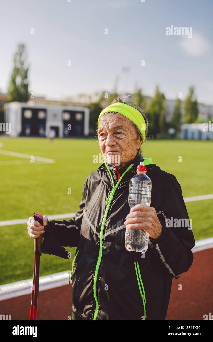 Seniorin übte nordic Walking im Stadion und hielt an, um Durst zu stillen, Wasser aus der Flasche zu trinken. Alter, Reife, aktiver Lebensstil und gut Stockfoto
