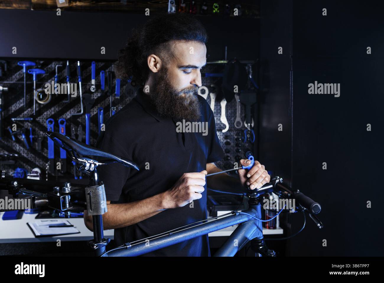 Thema Verkauf und Reparatur von Fahrrädern. Junge und stilvoll mit einen Bart und langes Haar, ein kaukasischer Mann benutzt ein Tool zur Einrichtung und ein Fahrrad in einem Speicher zu reparieren. B Stockfoto