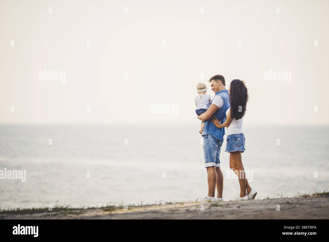 Familienurlaub in der Natur. Drei, Mutter, Vater, Tochter ein Jahr ständigen mit Rücken auf einer Klippe mit Blick auf das Meer. Mann hält Hild in Arme, Frau umarmen Hus Stockfoto