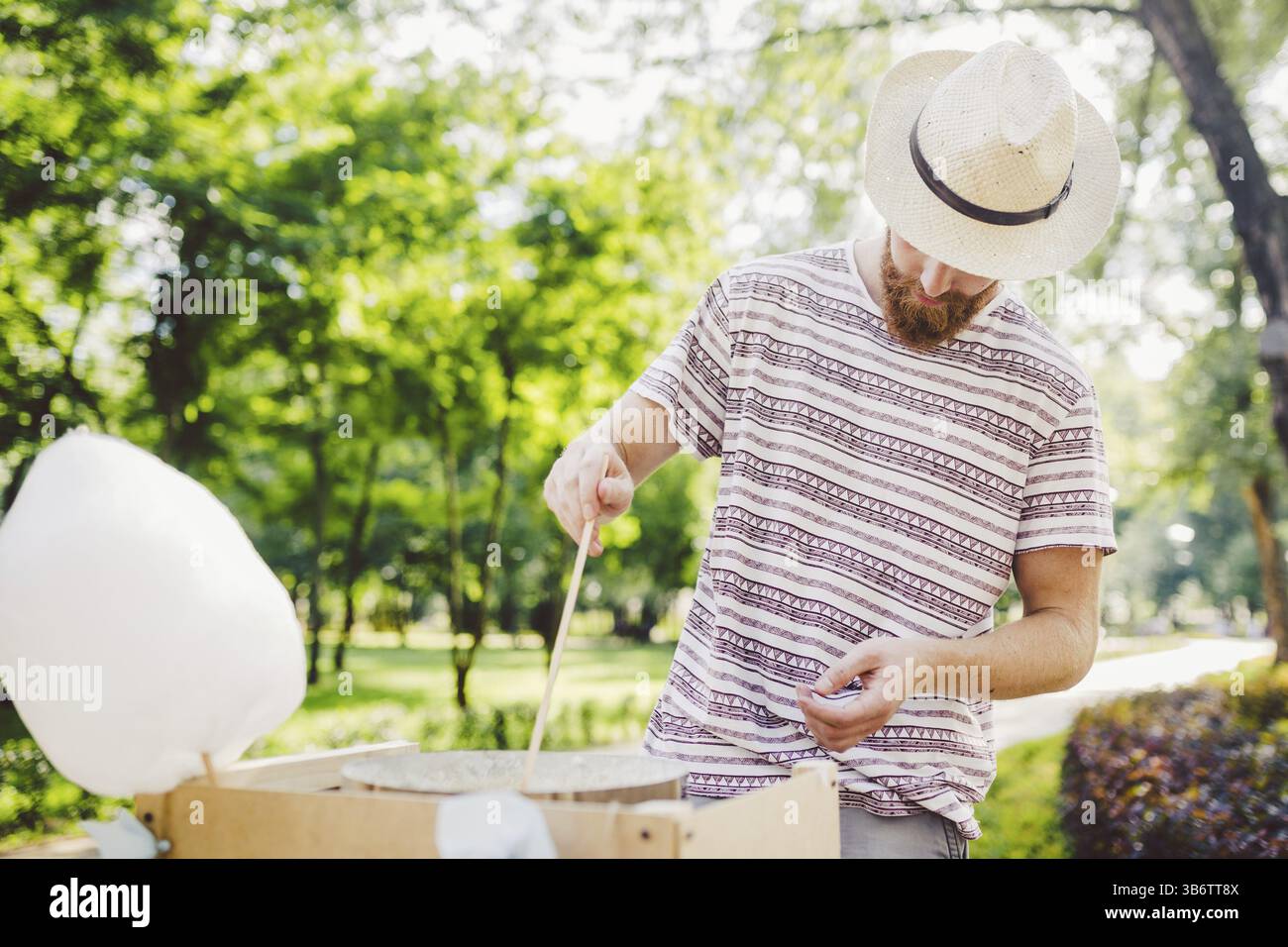 Fotomotiv für kleine Unternehmen, die Süßigkeiten kochen. Ein junger Mann mit dem Bart eines kaukasischen Händlers im Hut, der Besitzer des Outlets macht Zuckerwatte, Fee Stockfoto