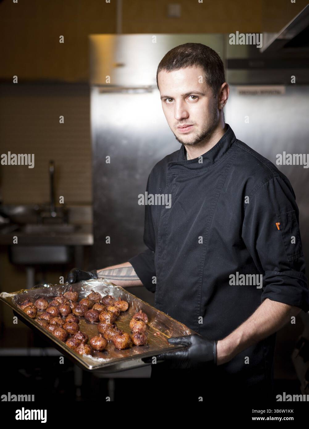 Portrait männlicher Koch mit gekochten Speisen in der Küche. Themenkochen. Junger kaukasischer Mann in schwarzer Uniform, Latexhandschuhe im Restaurant in der Küche. d Stockfoto