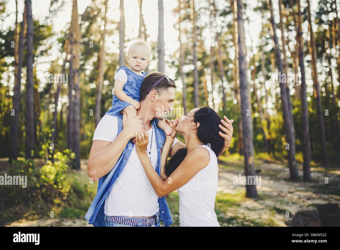 Familie Mama, Papa und Tochter sitzt bei Papa auf den Schultern, und Eltern Kuss auf die Natur im Wald im Sommer bei Sonnenuntergang Stockfoto