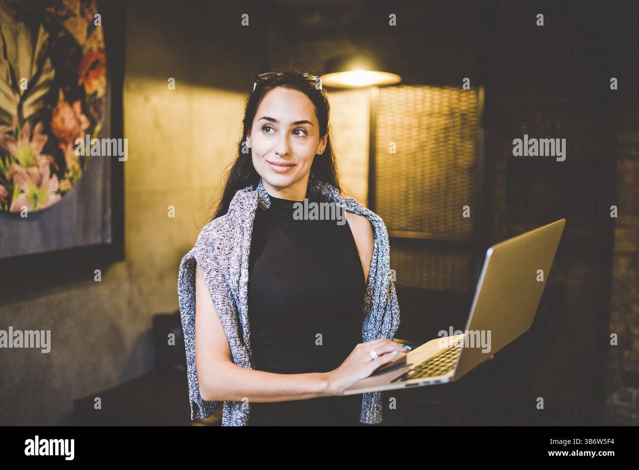 Das Konzept des Kleinunternehmens und der Technologie. Junge schöne Brünette Geschäftsfrau in schwarzem Kleid und grauem Pullover steht im Café mit Laptop Stockfoto