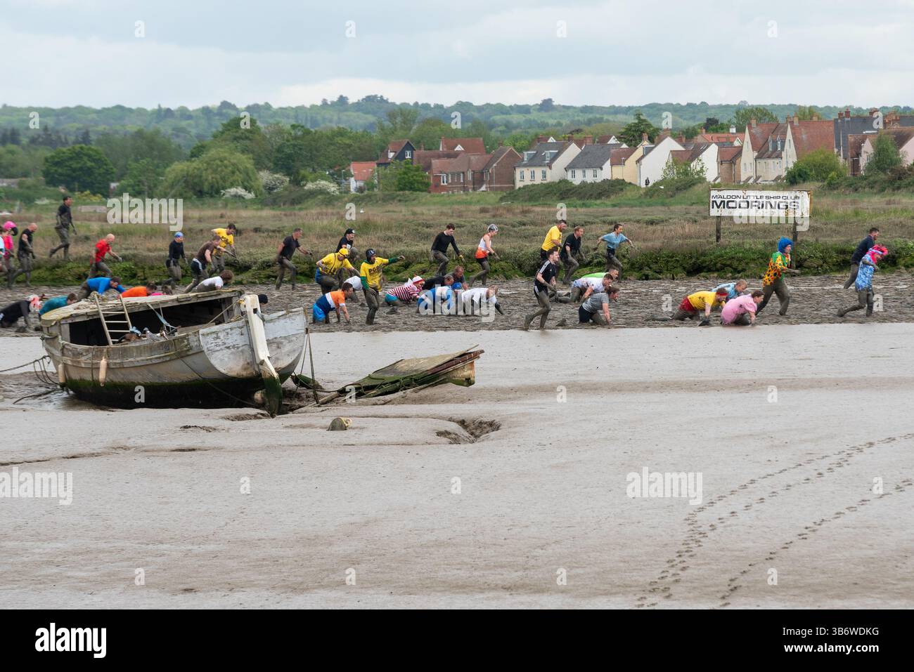 Promenade Park, Maldon, Essex, Großbritannien. Mai 2025. Eine große Anzahl von Läufern ging auf den zermürbenden Kurs, der sie bei Ebbe über den Fluss Chelmer und zurück führte, durch den anhaftenden, rutschigen Schlamm. Die Wettkämpfer des Maldon Mud Race treten für wohltätige Zwecke an. Viele tragen schicke Kleider und alle werden während des Rennens vom schleimigen Schlamm bedeckt, besonders diejenigen, die weiter hinten sind, wenn der Schlamm aufgewühlt ist. Teilnehmer, die an verfallendem Boot und Liegeplatz vorbeifahren Stockfoto