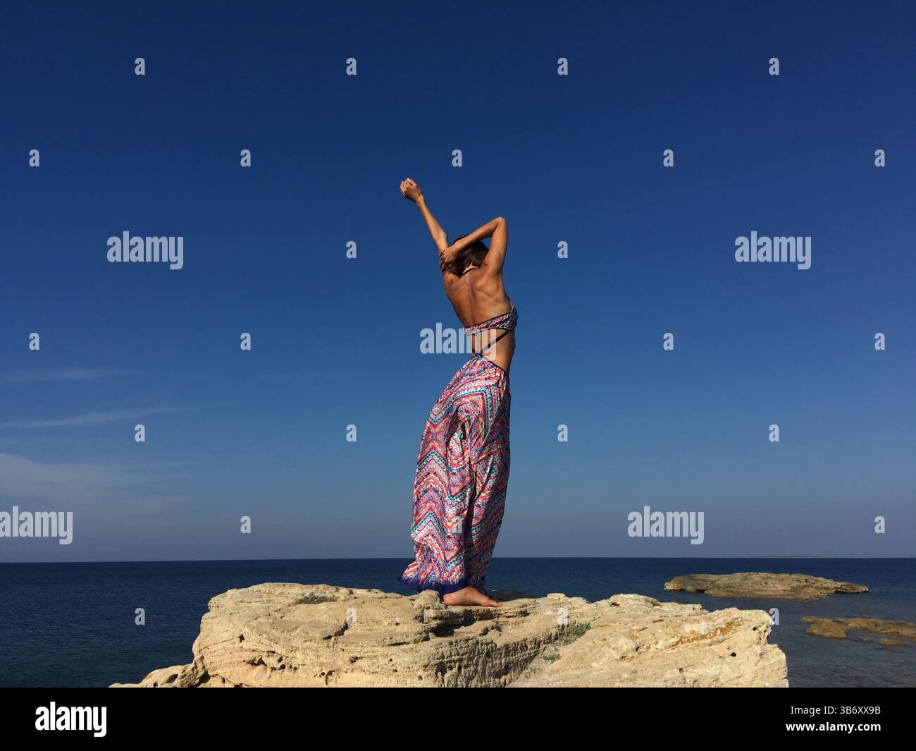 Schöne Frau in langem, farbenfrohen Rock, die am Rand der Klippe in Sardinien, Italien, mit erhobenen Armen wegblickt. Das Modemodell macht Spaß. Stockfoto