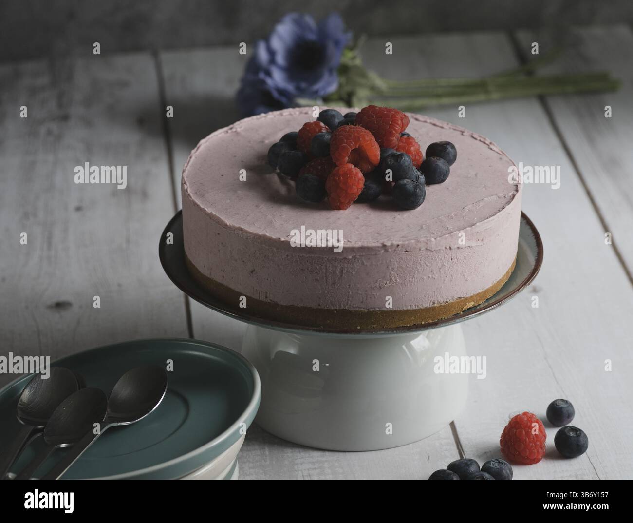 Horizontales Foto von einem gefrorenen roten Obstkuchen zusammen mit einigen Tellern, die auf einem weißen Holztisch in einer rustikalen und romantischen Umgebung serviert werden können. Stockfoto