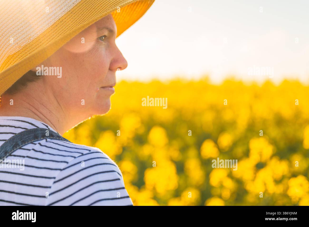 Rückansicht einer Landwirtin mit Strohhut, die auf blühendem Rapsfeld steht und in die Entfernung am Horizont blickt, selektiver Fokus Stockfoto