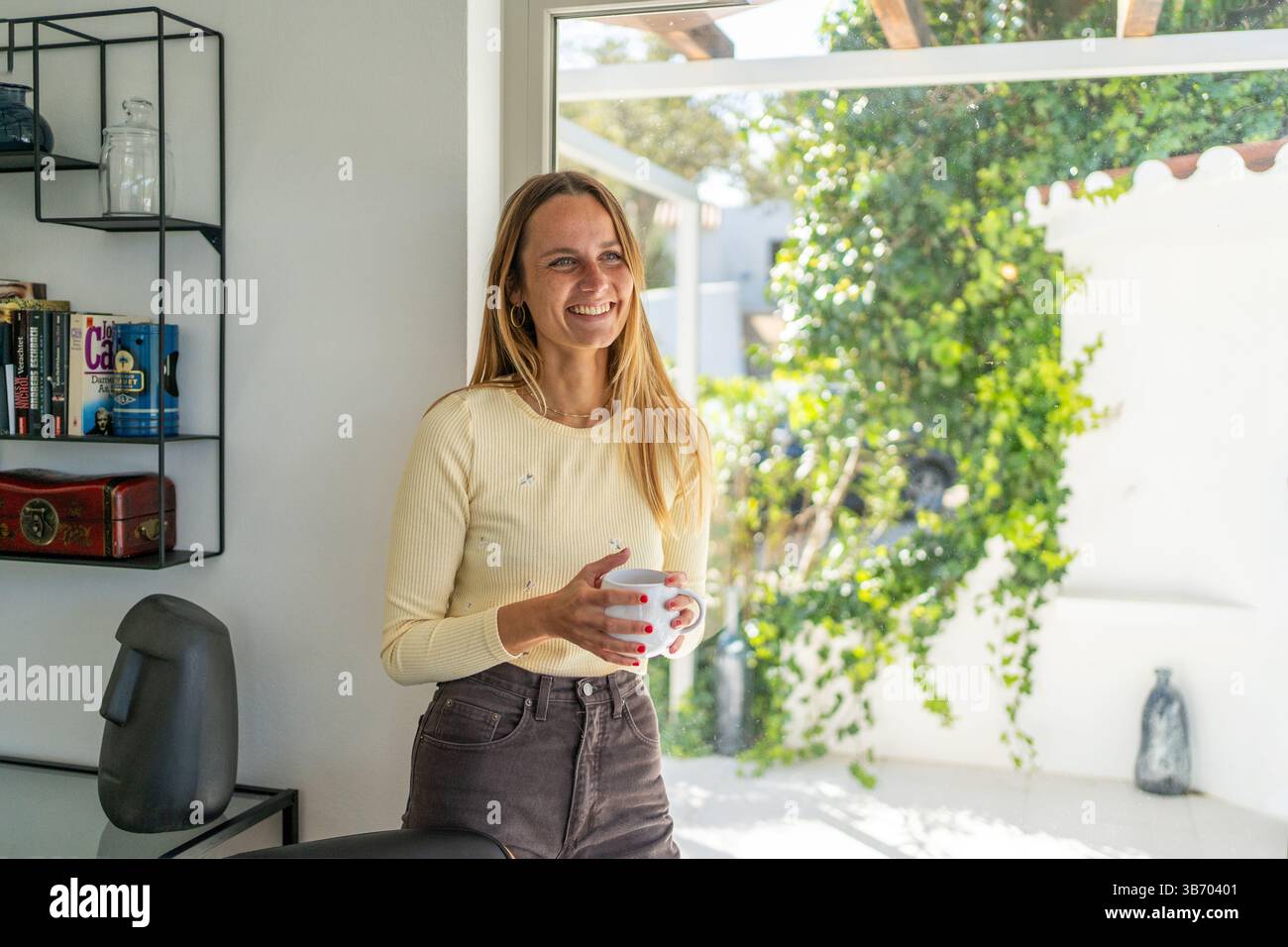 Frau zu Hause am Fenster, beginnt den neuen Tag, lacht mit Tee- oder Kaffeetasse auf der Hand. Wohlbefinden genießen, guten Morgen, glücklich, entspannend Stockfoto
