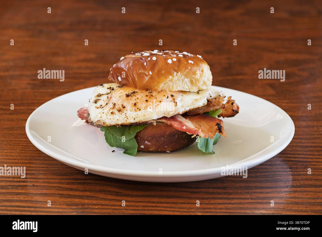 Sandwich mit Eierspeck und Rucola auf einem kleinen Brezelbrötchen und weißem Teller Stockfoto