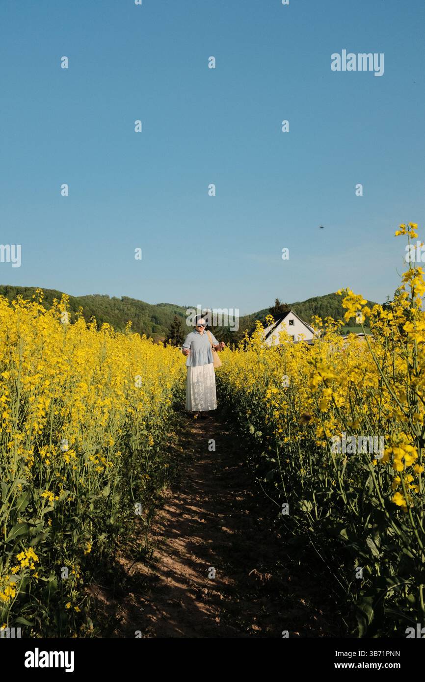 Frau in weißem Sommerkleid und Strohhut, die durch gelbes Rapsblumenfeld unter klarem blauem Himmel auf dem Land spaziert. Hochwertige Fotos Stockfoto