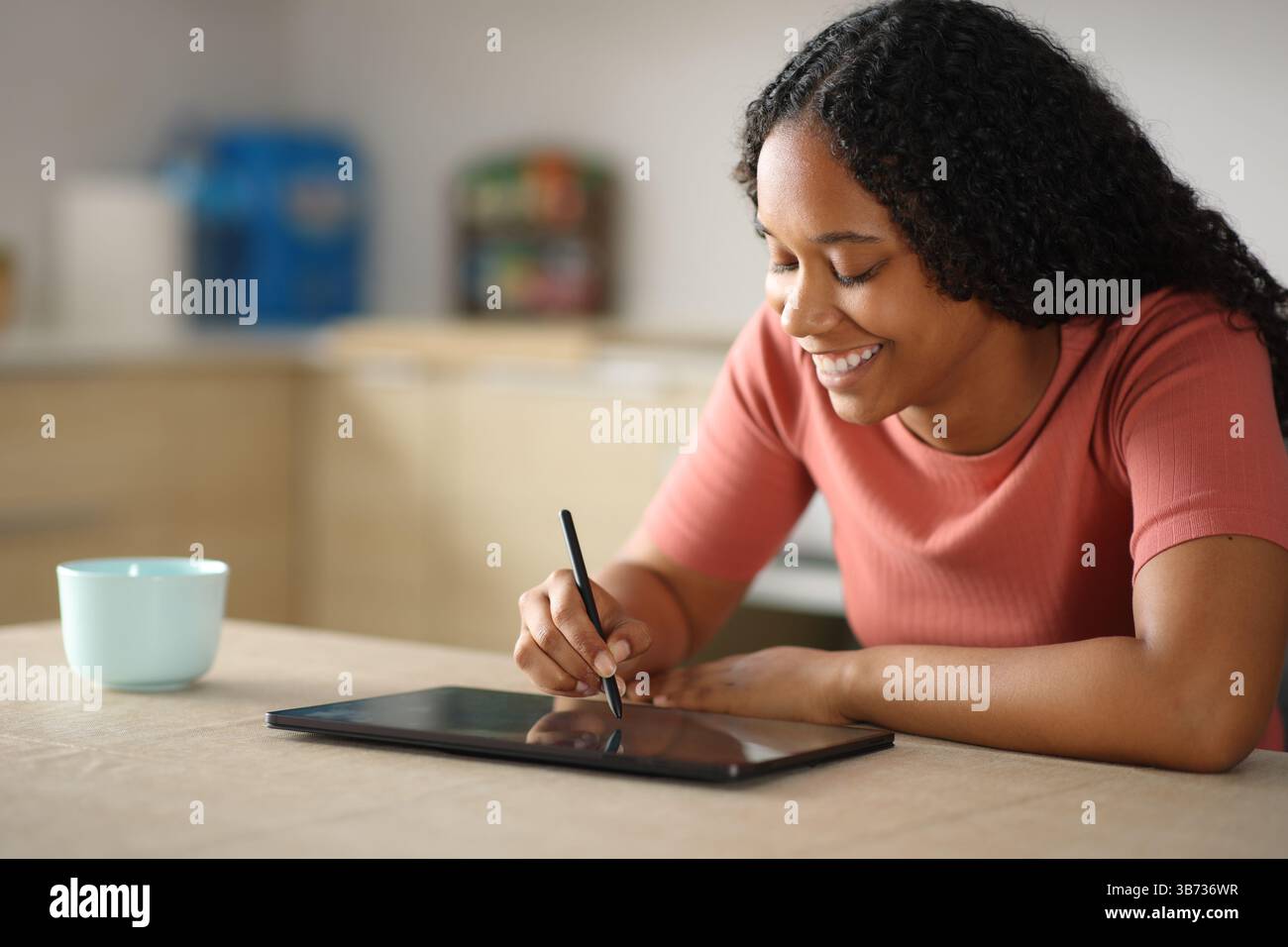 Glückliche schwarze Frau, die mit einem Stift in einem Tablet in der Küche zu Hause zeichnet Stockfoto