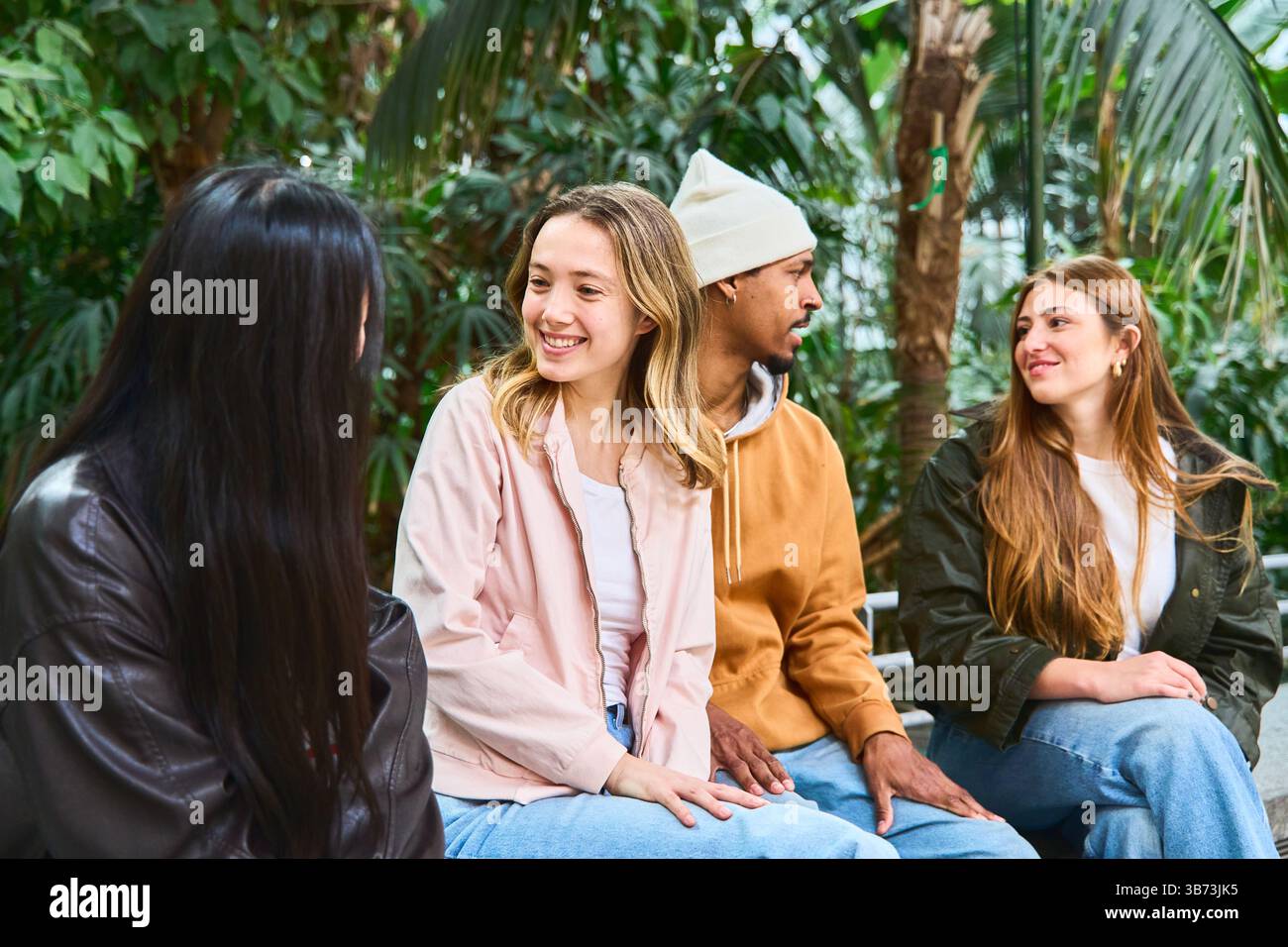 Vier Universitätsstudenten unterhalten sich gemeinsam, sitzen und entspannen in einem botanischen Garten voller üppiger Pflanzen Stockfoto