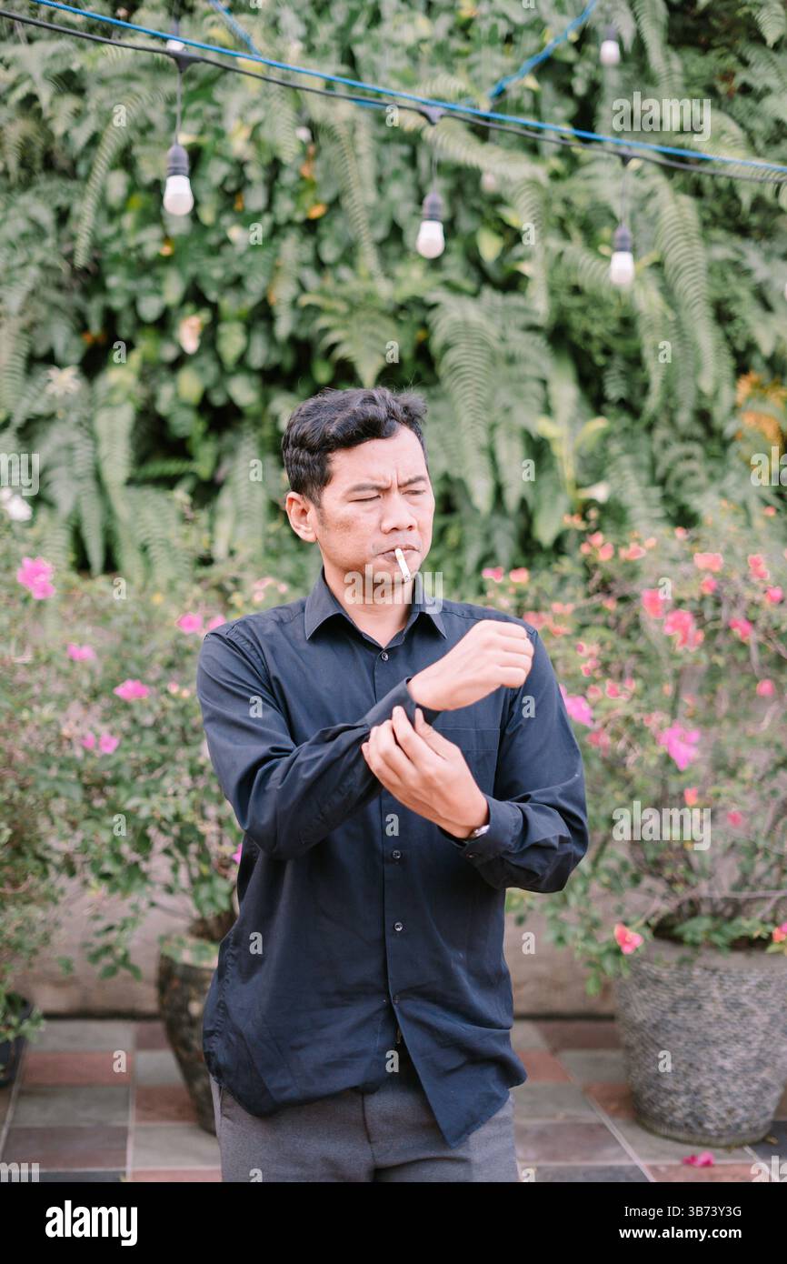 Ein asiatischer Mann in dunklem Hemd, der die Manschette anpasst, während er draußen zwischen Blumen und Grün raucht Stockfoto