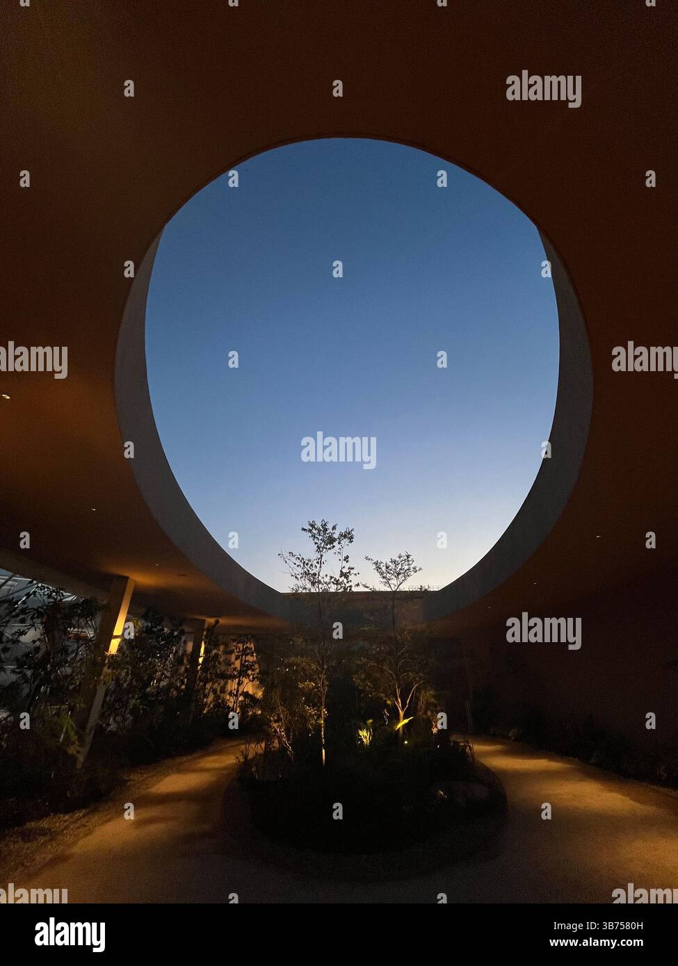 Die kreisförmige architektonische Öffnung umrahmt den Abendhimmel über einem minimalistischen Innenhof, der Natur und Design in einem ruhigen, modernen Raum vereint. - Smartphone-aufgenommenes Stockfoto