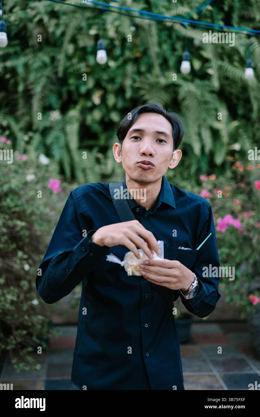 Ein junger Mann, der im Freien einen Snack genießt, umgeben von Grün und Umgebungslichtern, die eine ungezwungene und entspannte Atmosphäre ausstrahlen Stockfoto