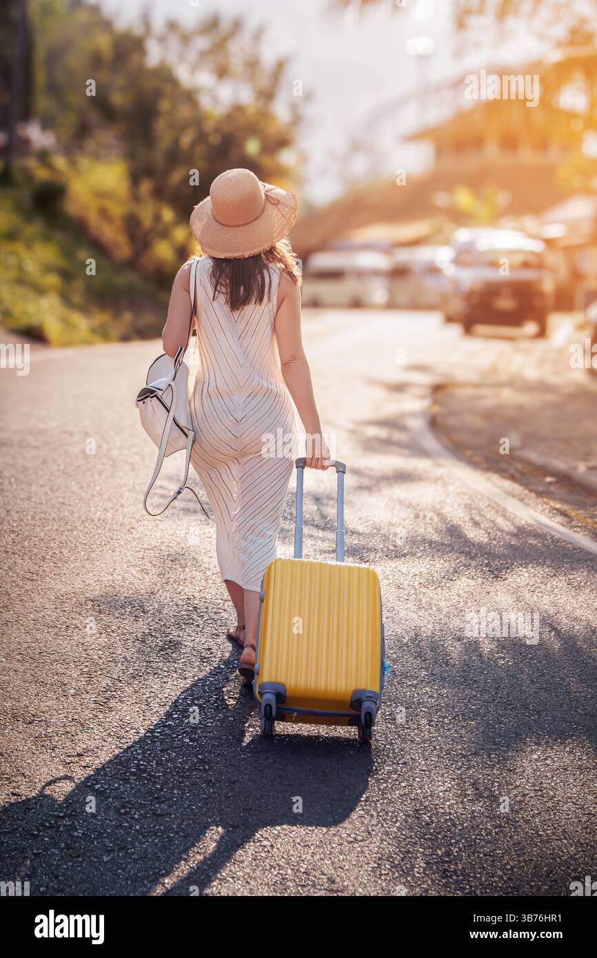 Frauen in Alleinreisen und Tourismus. Eine Reisende Frau mit breiter Krempe und Boho-Kleid läuft mit einem gelben Koffer auf einer gewundenen Straße entlang Stockfoto