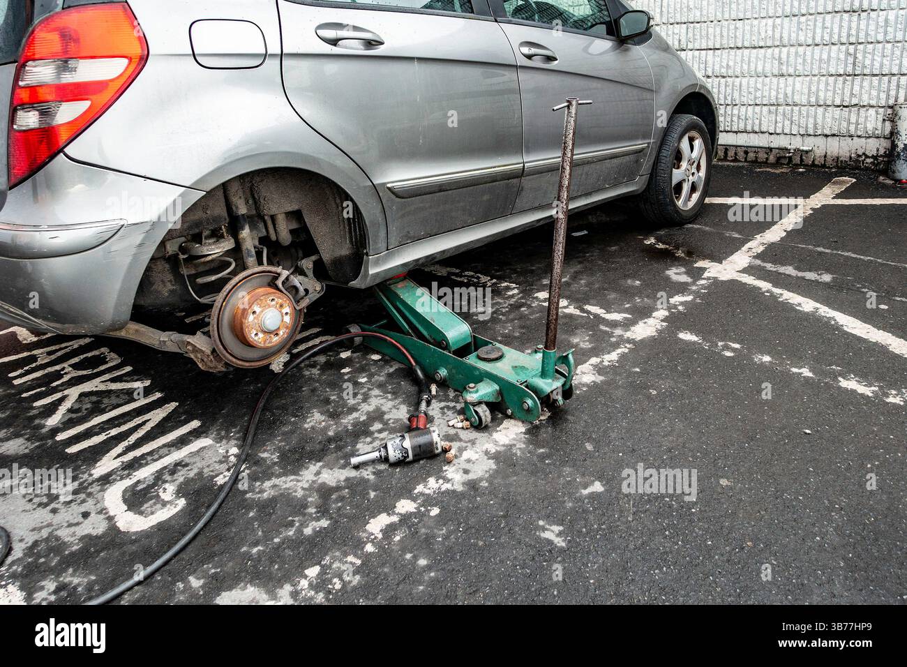 Ein silbernes Auto steht auf einem Parkplatz auf einem Wagenheber, ein Rad entfernt und Werkzeuge in der Nähe, um tagsüber einen Tiye-Wechsel durchzuführen. Stockfoto