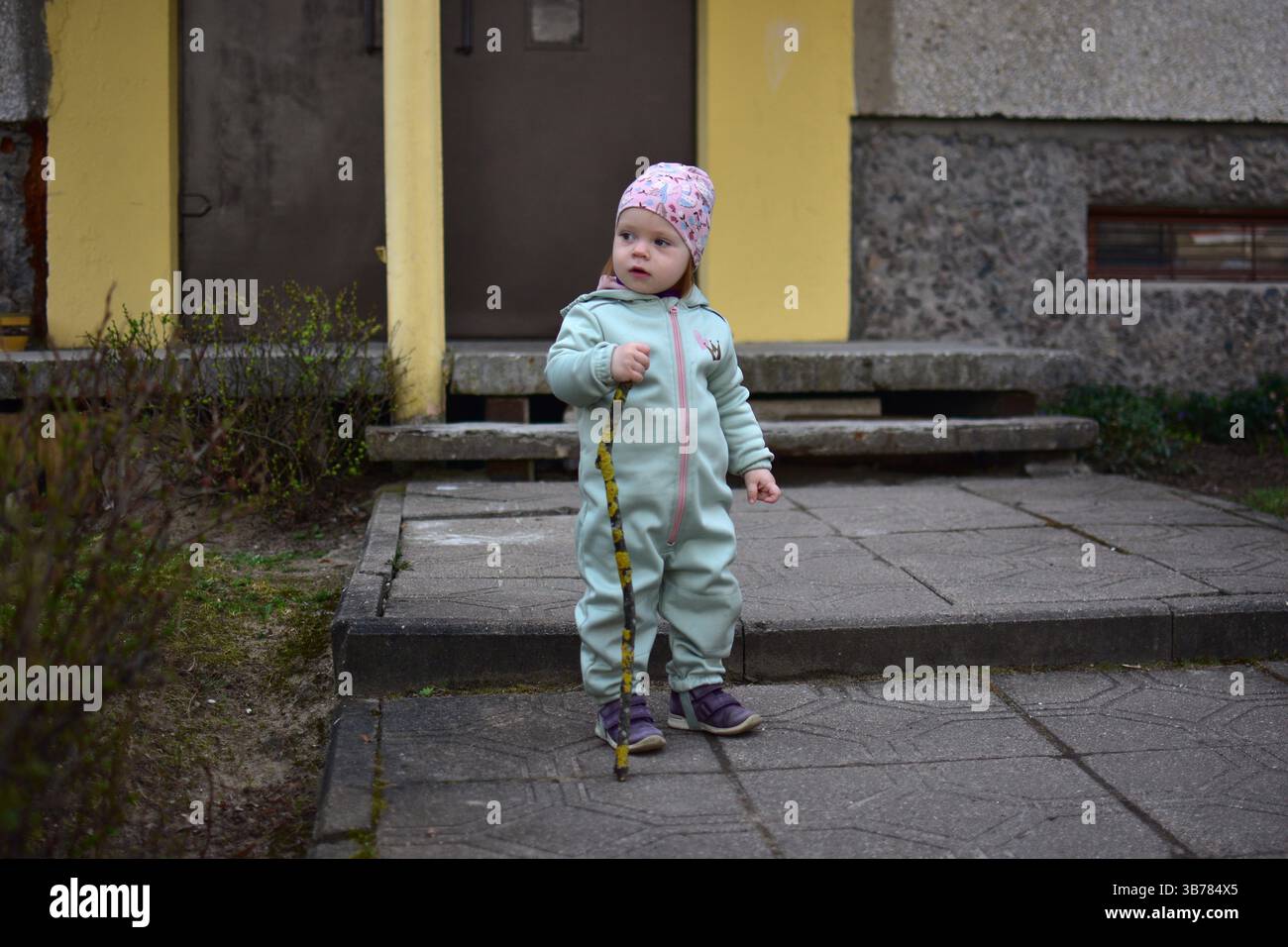 Baby Mädchen im mintfarbenen Softshell-Body hält einen Baumzweig und geht im Frühjahr neben dem Eingang zum Appartementhaus in Osteuropa. Kleinkinder Stockfoto