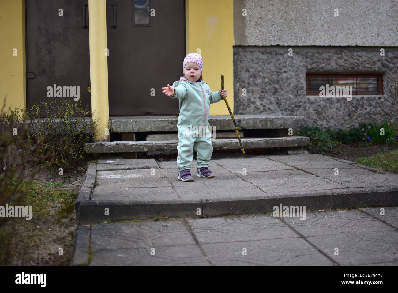Baby Mädchen im mintfarbenen Softshell-Body hält einen Baumzweig und geht im Frühjahr neben dem Eingang zum Appartementhaus in Osteuropa. Kleinkinder Stockfoto