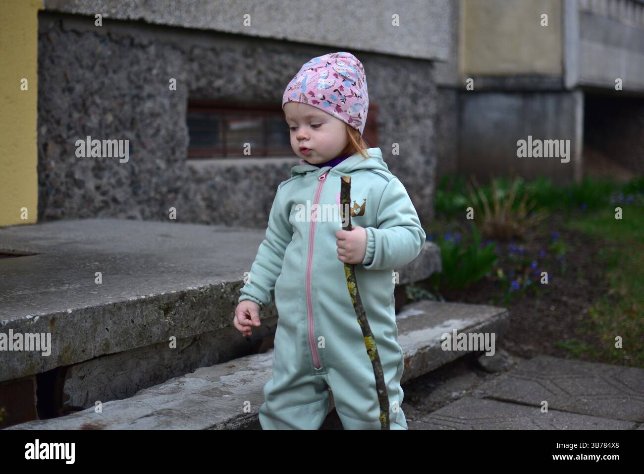 Baby Mädchen im mintfarbenen Softshell-Body hält einen Baumzweig und geht im Frühjahr neben dem Eingang zum Appartementhaus in Osteuropa. Kleinkinder Stockfoto