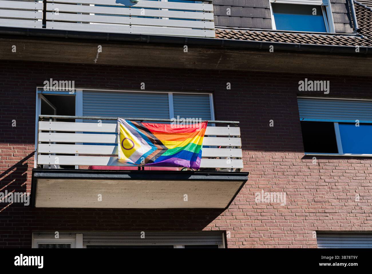Auf dem Balkon eines Wohngebietes hängt eine leuchtende Flagge mit Regenbogenfarben und zusätzlichen Symbolen. Die Sonne strahlt ein warmes Licht an Stockfoto