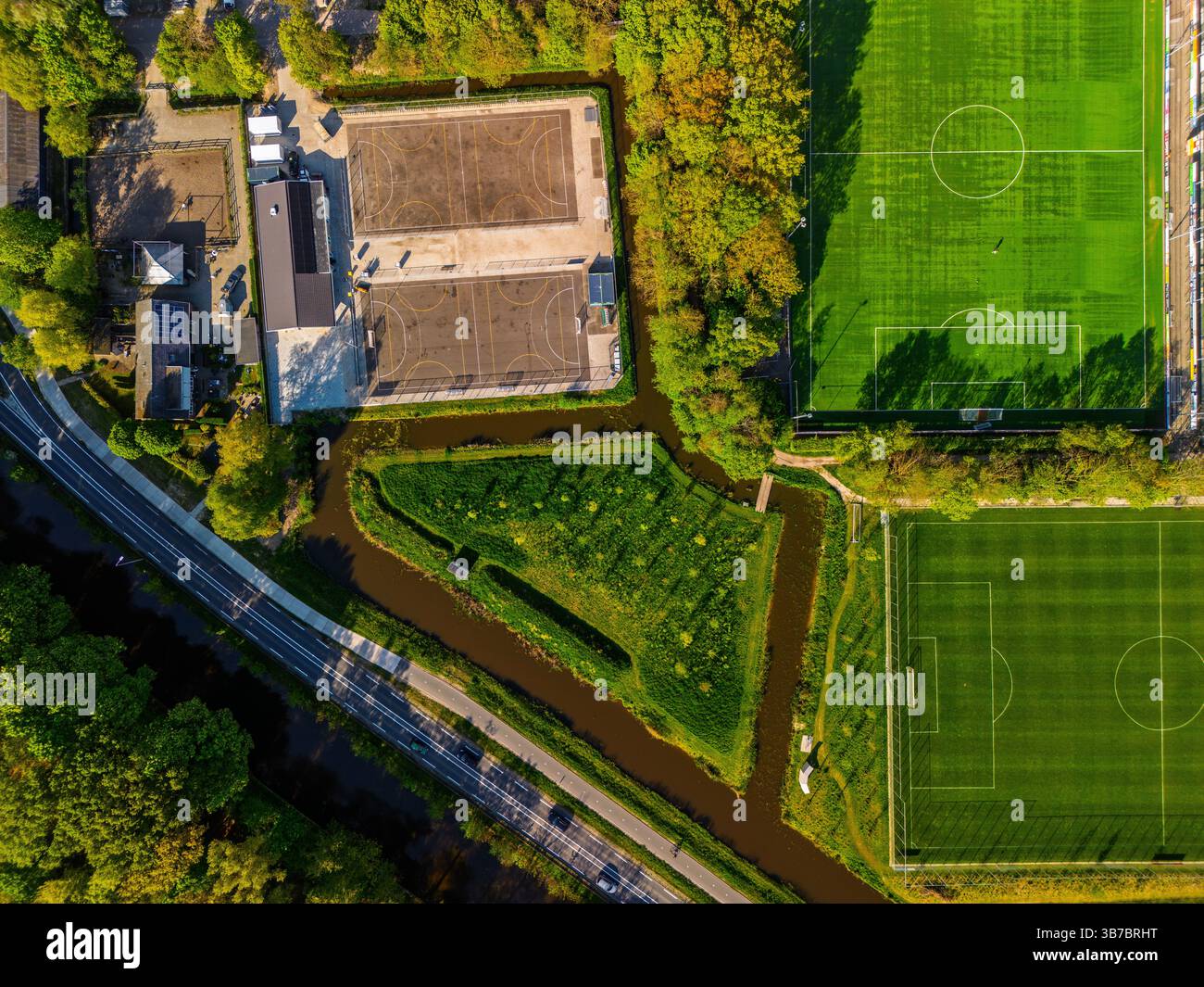Aus der Vogelperspektive auf einem grünen Sportplatz mit weißen Markierungen, umgeben von Bäumen und Fußwegen, mit hellem Sonnenlicht, das lange Schatten wirft. Hallo Stockfoto